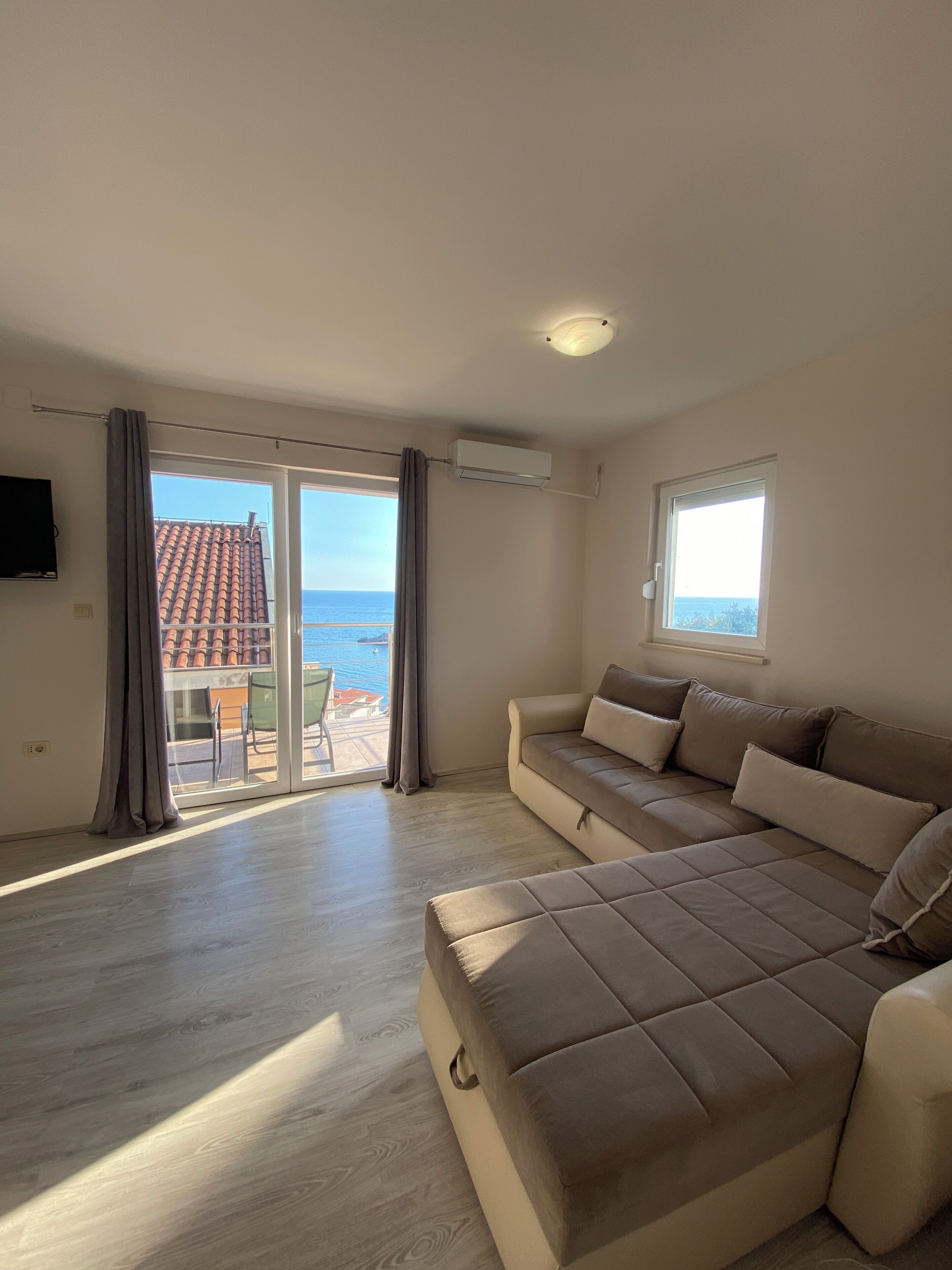 A modern living room with a beige sofa, a flat-screen TV, and a large sliding glass door leading to a balcony with a view of the sea.