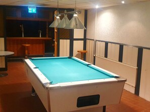 Une table de billard recouverte d’un drap bleu, une lampe suspendue et un coin bar à l’arrière-plan.