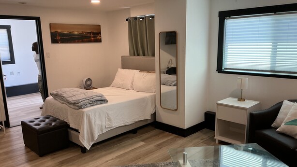 A modern bedroom with a bed, a glass coffee table, a nightstand, a mirror, and a window with blinds.
