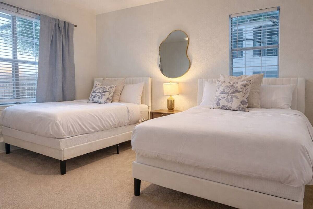 A hotel room with two beds, a large mirror, a nightstand with a lamp, and a window with blinds.
