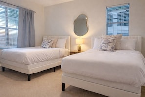 A hotel room with two beds, a large mirror, a nightstand with a lamp, and a window with blinds.