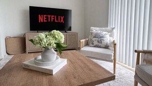A living room with a flat-screen TV displaying 'NETFLIX,' a white vase with flowers on a wooden coffee table, a beige armchair, and a light-colored sofa.
