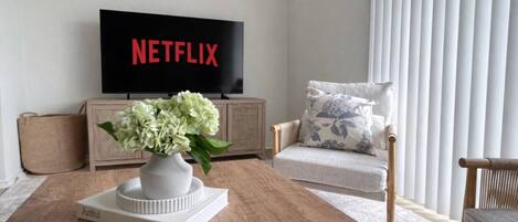 A living room with a flat-screen TV displaying 'NETFLIX,' a white vase with flowers on a wooden coffee table, a beige armchair, and a light-colored sofa.
