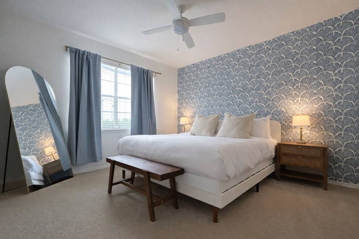 A bedroom with a bed, bedside tables, a bench, a ceiling fan, and a window with curtains.