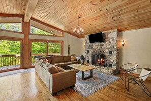 Ein gemütliches Wohnzimmer mit Kamin aus Stein, Holzboden und großen Fenstern mit Blick auf Grünflächen.