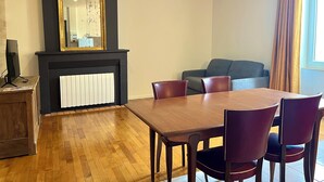 A dining area with a wooden table and chairs, a fireplace with a white radiator, a large mirror, and a sofa in the background.