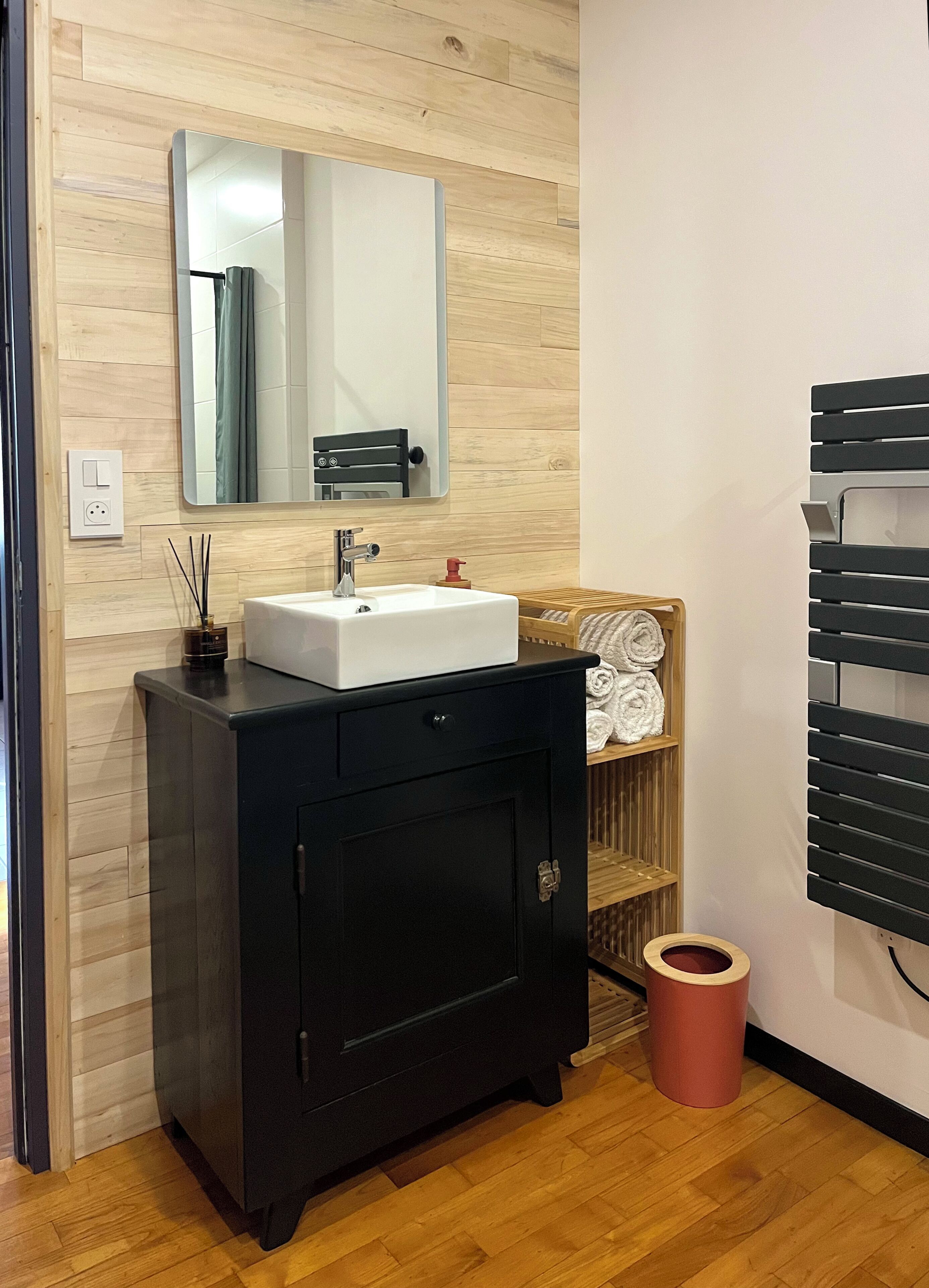 Une salle de bain avec un meuble-lavabo en bois, un lavabo blanc, un miroir, un porte-serviettes et une poubelle.