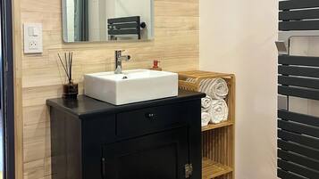 Un baño con tocador de madera, un lavamanos blanco, un espejo, un perchero para toallas y un cubo de basura.