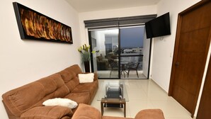A living room with a brown sofa, a glass coffee table, a flat-screen TV mounted on the wall, and a sliding glass door leading to an outdoor balcony. - Bonito departamento con vista al mar  (Mazatlán)