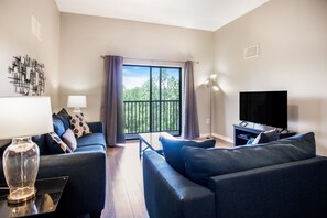 Ein modernes Wohnzimmer mit einer Couch, einem Fernseher, einer Lampe und Blick auf Bäume durch ein großes Fenster.