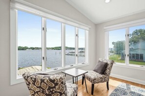 Ein Zimmer mit großen Fenstern, einem gemusterten Sessel, einem kleinen Tisch und Blick auf ein Gewässer.