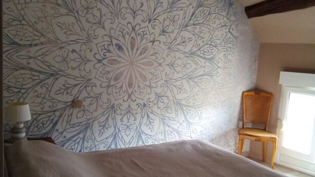 A bedroom with a large patterned wall, a bed with a beige blanket, a wooden chair, and a window. - Champagne character apartment (Essoyes)