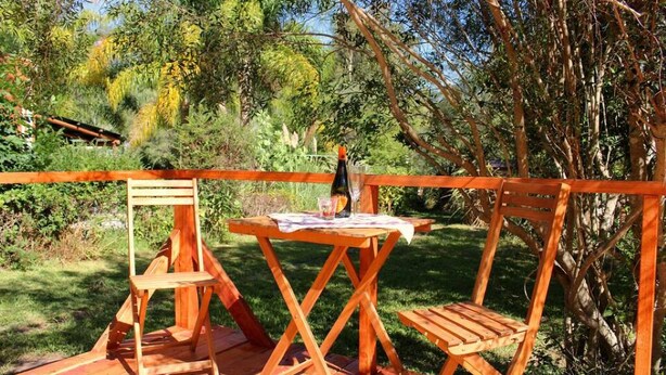 Una terraza de madera al aire libre con dos sillas y una mesa, rodeada de follaje y árboles.