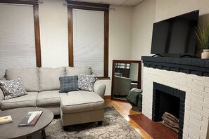 A living room with a sofa, ottoman, fireplace, and a flat-screen TV. - Cozy Updated 2BR Home Near Carondelet Park (St. Louis)