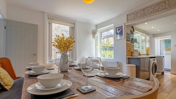 Un comedor con una mesa de madera dispuesta para cuatro personas, un jarrón con flores y una cocina al fondo.