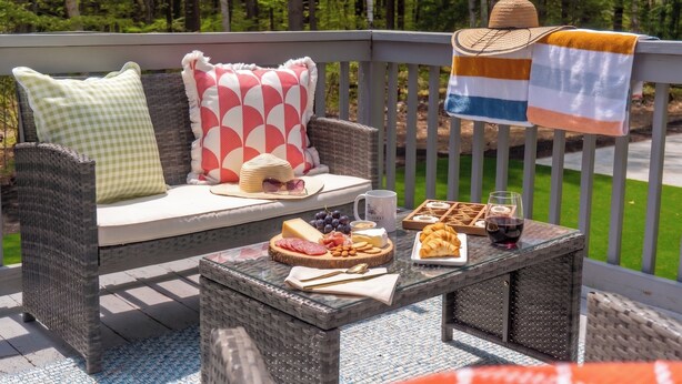 A wicker sofa with patterned cushions, a glass table with food and drink, and a view of a forested area. - Wells Retreat: 3BR, Firepit, Pickleball, Sleeps 9 (Wells)
