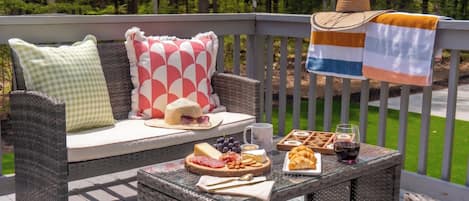 Un canapé en osier agrémenté de coussins à motifs, une table en verre avec de la nourriture et des boissons, et une vue sur une zone boisée.