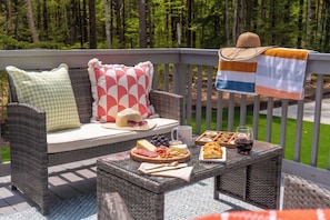 Un canapé en osier agrémenté de coussins à motifs, une table en verre avec de la nourriture et des boissons, et une vue sur une zone boisée.
