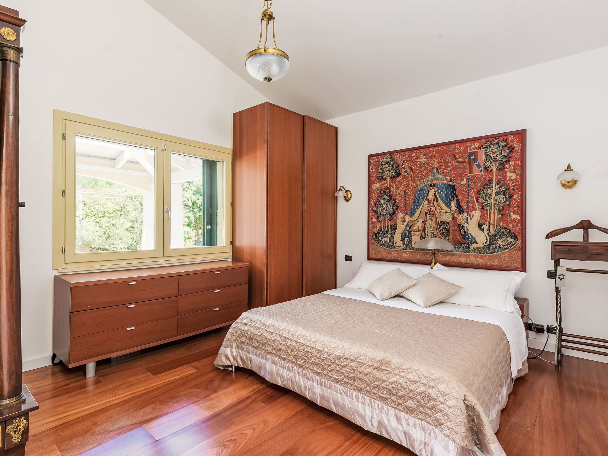 Un dormitorio con cama, armario, ventana, cuadro y mesita de noche.