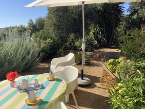 Un espace de restauration en plein air avec une table dressée pour deux personnes, entouré de verdure et surplombé d’un parasol blanc.
