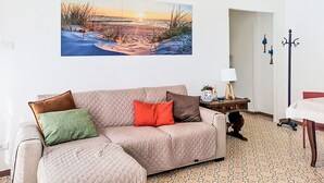 Ein Wohnzimmer mit einem großen Gemälde einer Strandansicht, einem beigefarbenen Sofa mit Kissen, einem kleinen Tisch mit einer Lampe und einem gemusterten Teppich.