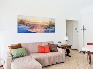 A living room with a large painting of a beach scene, a beige sofa with pillows, a small table with a lamp, and a patterned carpet. - Bemi' al mare by Interhome (Terracina)