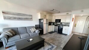 A modern living room with a grey sofa, a black coffee table, and a kitchen area with white cabinets and a black refrigerator. - Sun Filled Corner Unit w/ Gulf Views in Every Room, Steps Away from Babe's Beach (Galveston)
