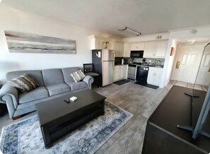 Ein modernes Wohnzimmer mit grauem Sofa, einem schwarzen Couchtisch und einer Küchenzeile mit weißen Schränken und einem schwarzen Kühlschrank.