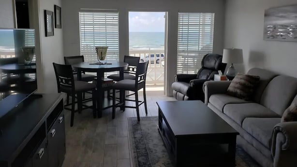 A living room with a balcony, a dining table with chairs, a sofa, a TV, and a lamp. - Sun Filled Corner Unit w/ Gulf Views in Every Room, Steps Away from Babe's Beach (Galveston)
