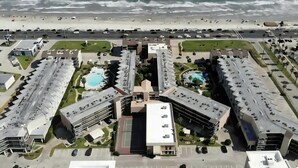 An aerial view of a coastal hotel with multiple buildings, a central pool, and a beach in the background. - Sun Filled Corner Unit w/ Gulf Views in Every Room, Steps Away from Babe's Beach (Galveston)