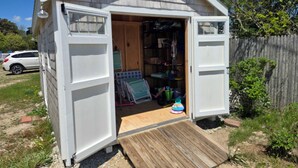 A small shed with an open door, revealing storage shelves and a wooden ramp leading to the entrance.