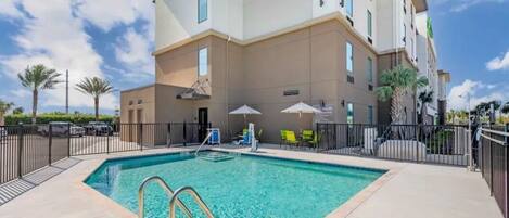 A hotel with a swimming pool, outdoor seating, and a clear sky.