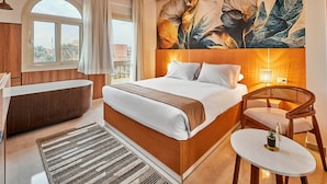 A hotel room with a large bed, a bathtub, a chair, a small table, and a large window.