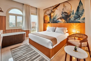 A hotel room with a large bed, a bathtub, a chair, a small table, and a large window.