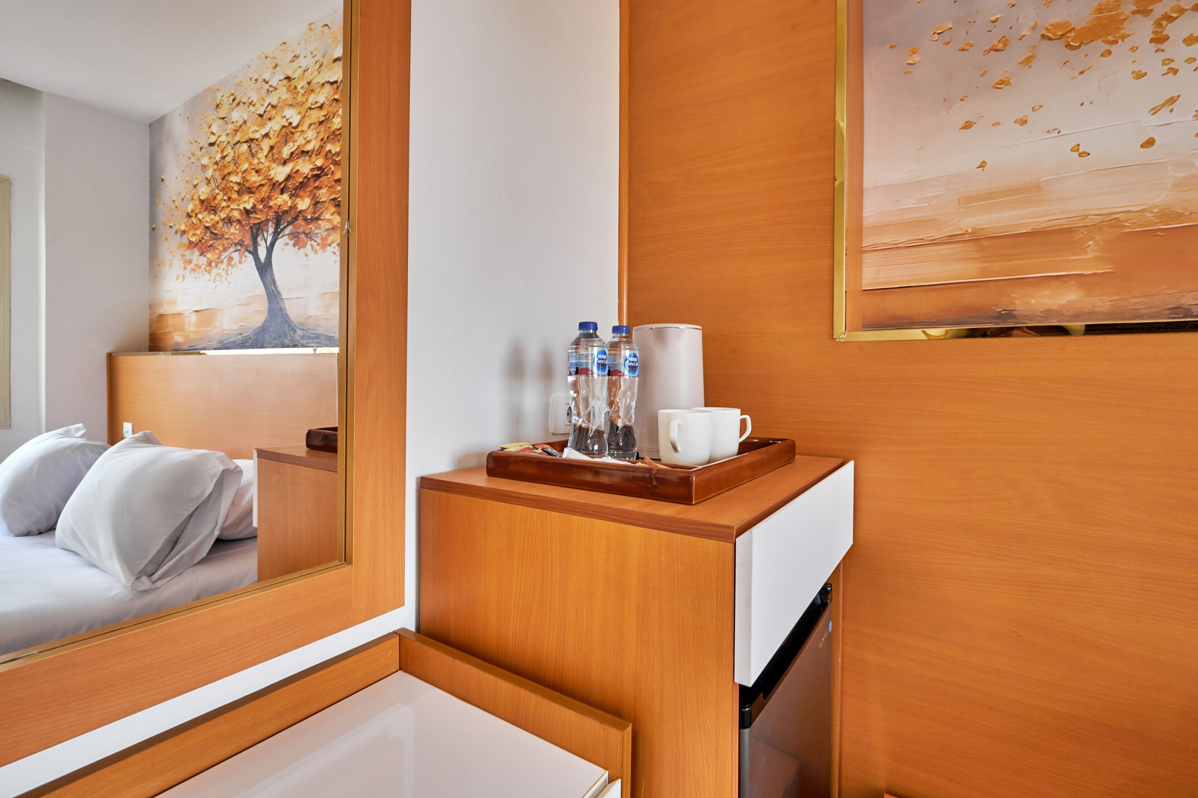 Een hotelkamer met een bed, een houten nachtkastje met een dienblad met waterflesjes en een beker, en een schilderij van een boom aan de muur.