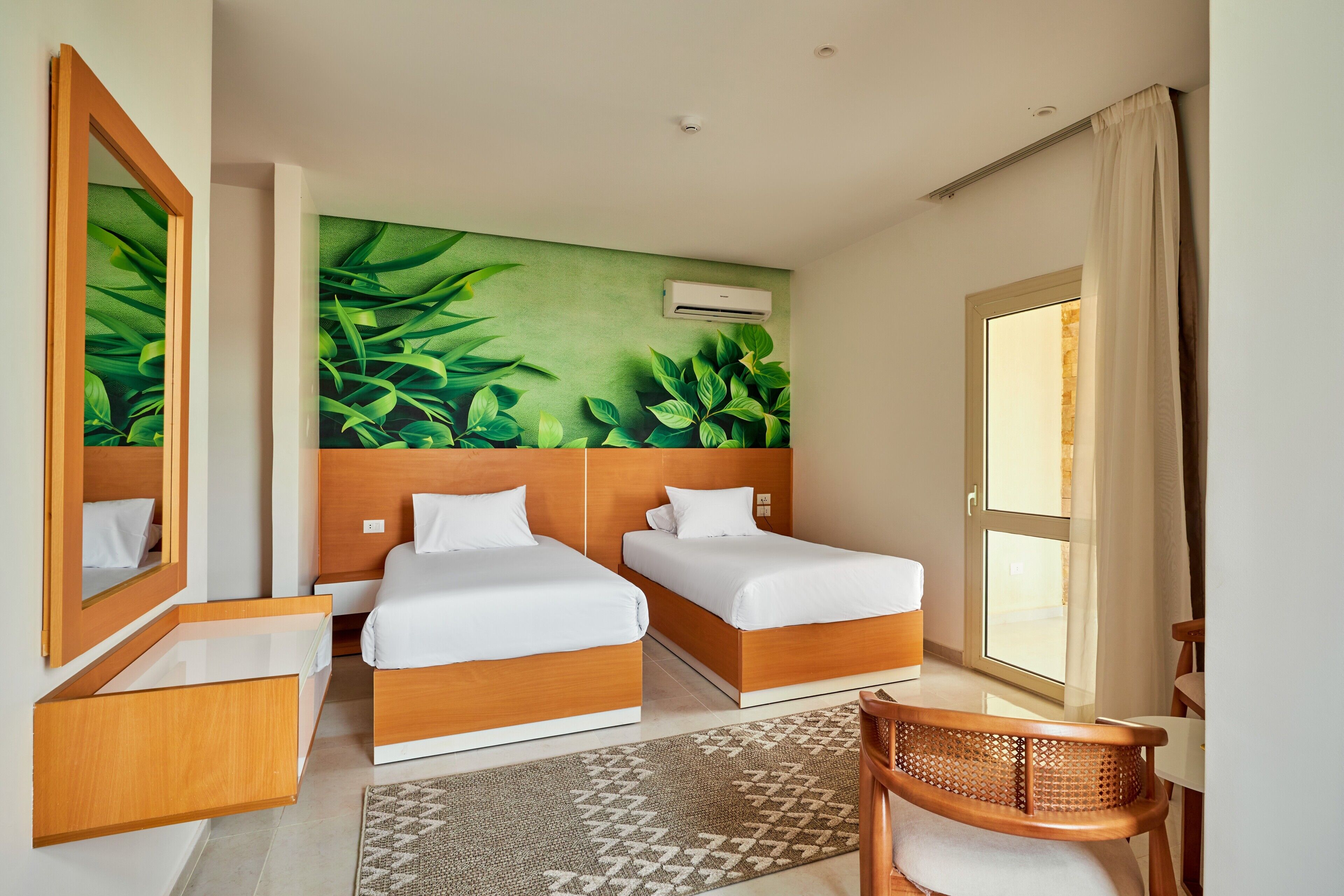 A hotel room with two beds, a large window, a mirror, a wall-mounted air conditioner, and a patterned rug.