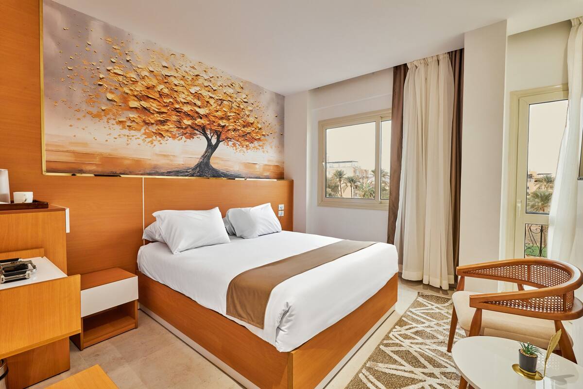 A hotel room with a bed, a desk, a chair, a painting of a tree, and a view of the outside.