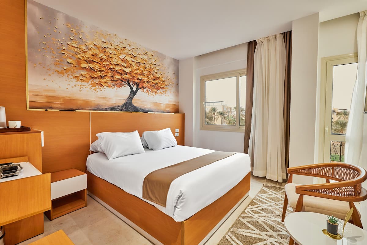A hotel room with a bed, a desk, a chair, a painting of a tree, and a view of the outside.