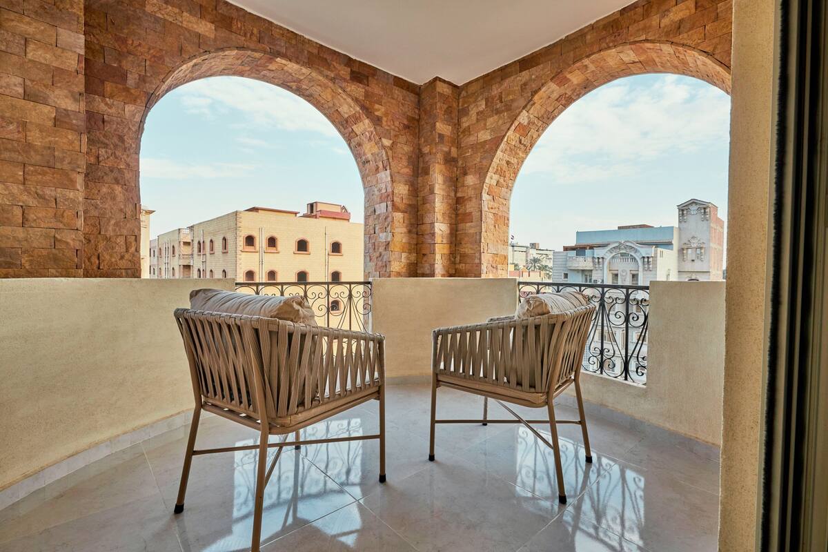 A balcony with two wicker chairs, a wrought iron railing, and a view of buildings through arched brick walls.
