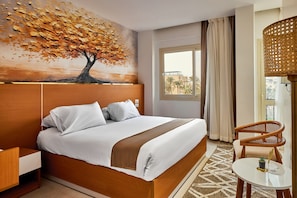 A hotel room with a large bed, a nightstand, a chair, a small table, and a window with curtains. - PALMERAS PYRAMIDS HOTEL (Giza)