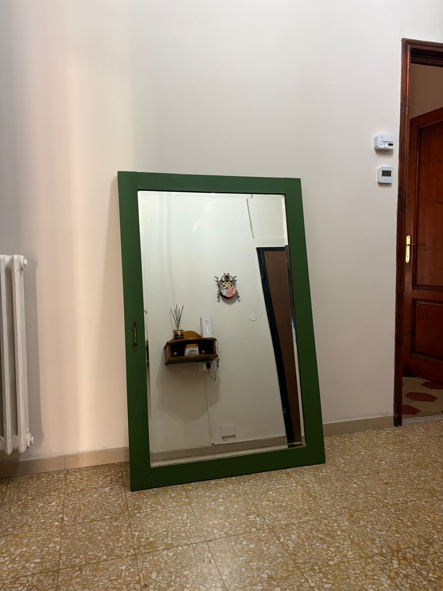Una habitación con un espejo de marco verde, un radiador, un estante pequeño con adornos y una puerta de madera.