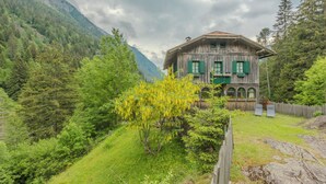 A rustic wooden house with green shutters, surrounded by lush greenery and mountains.