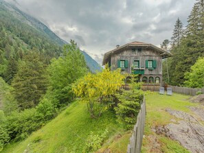 A rustic wooden house with green shutters, surrounded by lush greenery and mountains.