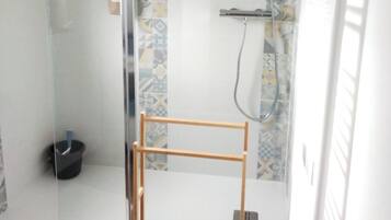 A glass-enclosed shower with a tiled wall, a wooden rack, and a black bucket.