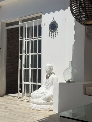 Eine weiße Buddha-Statue, eine Glasvase und ein Wandbehang vor einem Gebäude mit einer steinernen Fassade und einer weißen Tür.