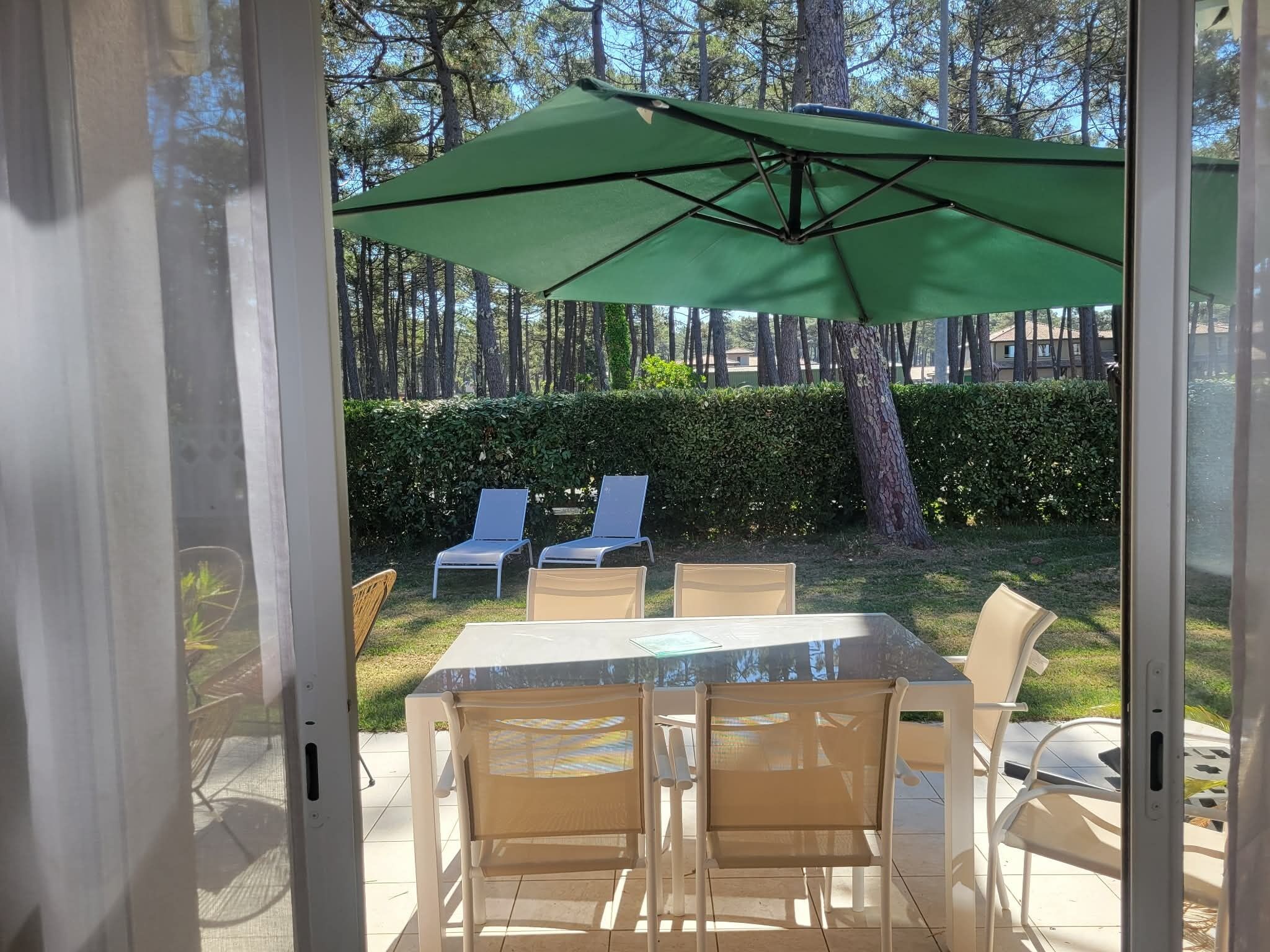En udendørs terrasse med et bord og stole, en grøn parasol samt udsigt til en hæk og træer.
