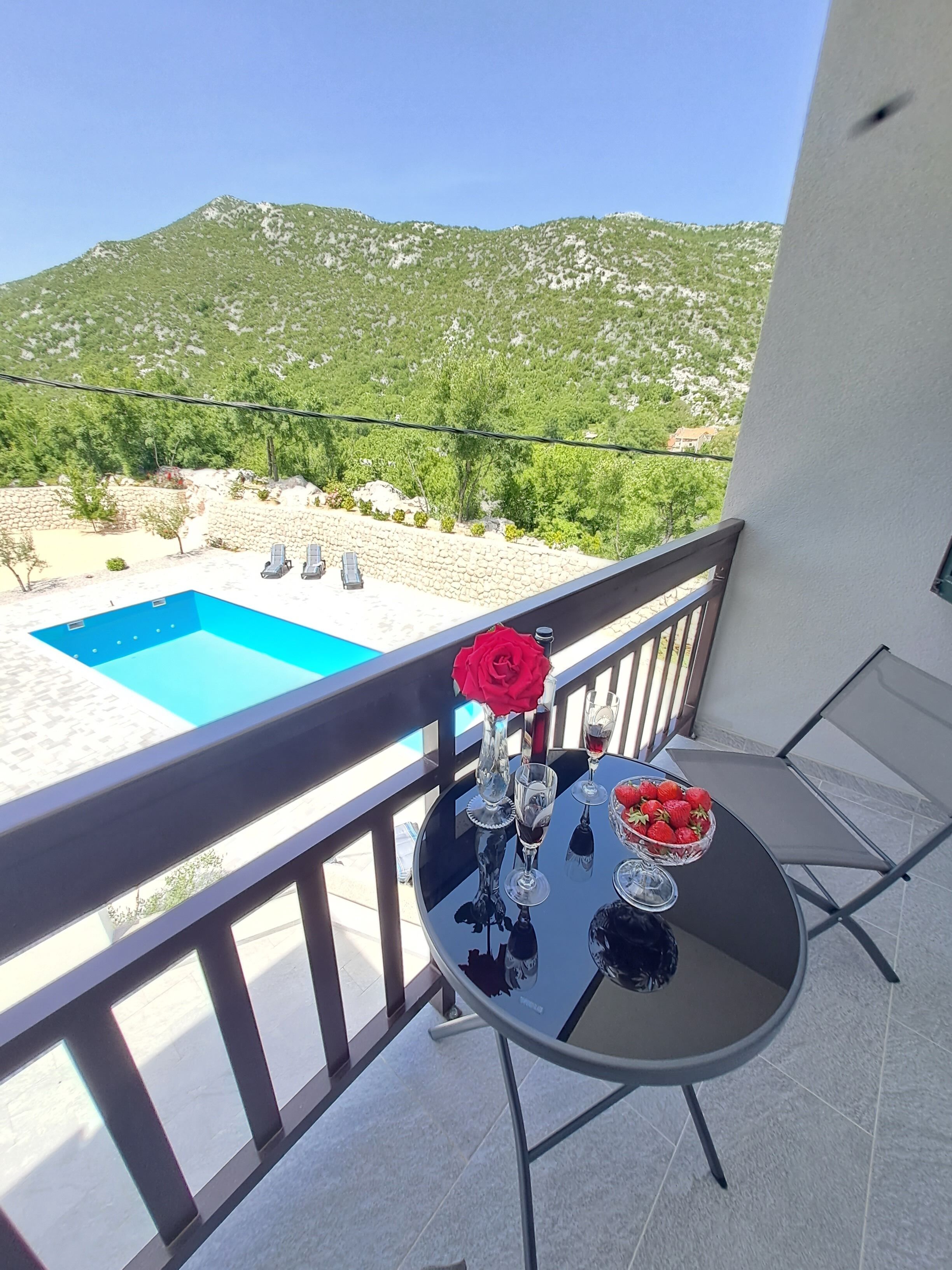 A balcony with a table, chairs, a vase with a red rose, a bowl of strawberries, and a view of a pool and mountains.