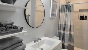 Un baño con lavamanos blanco, espejo y retrete. Hay toallas apiladas en un estante, y una cortina de ducha con estampado a rayas.