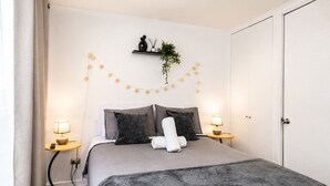 A neatly arranged bedroom with a bed, bedside lamps, a shelf with a plant, and a string of lights. - San Isidro 635 (Santiago)