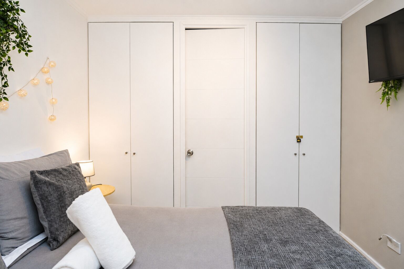 Un dormitorio con cama, mesita de noche, lámpara y televisor montado en la pared.